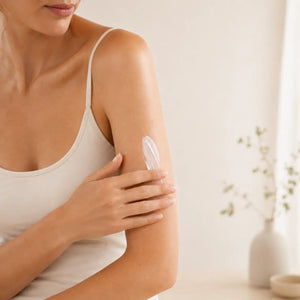 A woman wearing a white tank top is applying lotion to her arm.